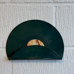 Könnte beinhalten: Ein dunkelgrüner Vinyl-Schallplattenhalter an einer weißen Backsteinmauer. Der Halter zeigt eine Vinyl-Schallplatte und zeigt ihr Etikett. Das halbkreisförmige Design des Halters bietet eine einzigartige Möglichkeit, Schallplatten zu präsentieren.