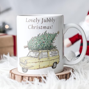 May include: White ceramic mug with the text "Lovely Jubbly Christmas!" and an illustration of a yellow van carrying a Christmas tree. The mug is on a wooden coaster, with a Santa Claus figure in the background.
