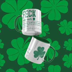 May include: Two white mugs with green trim. One mug has the word "FECK" in green letters and a definition of the word. The other mug has a green shamrock. Both mugs are against a green background with shamrock patterns.