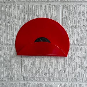 May include: A bright red vinyl record holder mounted on a textured white brick wall. The holder is designed to cradle a vinyl record, showcasing its vibrant color and design. The record itself is partially visible, adding a touch of nostalgia.