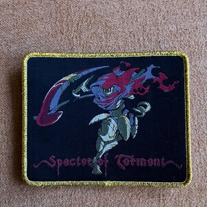 May include: A black embroidered patch with a gold border. The patch features a red and blue creature with a sword and the text "Specter of Torment".