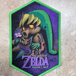 May include: A green, purple, and black embroidered patch featuring Link from The Legend of Zelda: Majora's Mask. The patch is shaped like a hexagon and has a green border.