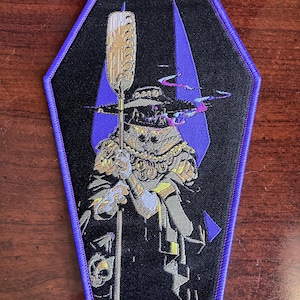 Könnte beinhalten: Ein gestickter Patch in Lila und Schwarz mit einer skelettartigen Figur, die einen großen Hut trägt und einen Stab hält. Die Figur steht vor einem lila und schwarzen Hintergrund in Form eines Sarges.