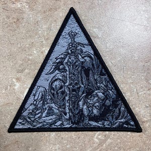 Puede incluir: Un parche triangular con borde negro presenta una detallada ilustración en blanco y negro de una figura esquelética sosteniendo una gran espada. El diseño tiene una apariencia texturizada, con sombreado y detalles intrincados. El parche es probablemente para ropa o accesorios.