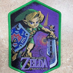 May include: A green, purple, and blue embroidered patch featuring Link from The Legend of Zelda: Majora's Mask. Link is holding a sword and shield, and the text "The Legend of Zelda: Majora's Mask" is visible at the bottom.