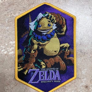 May include: A purple and gold embroidered patch featuring a character from The Legend of Zelda: Majora's Mask. The character is a large, yellow, frog-like creature with a blue necklace and a purple background.