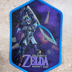 May include: A blue and purple embroidered patch featuring Link from The Legend of Zelda: Majora's Mask. The patch shows Link holding his sword and standing in front of a purple background with a large moon in the background.