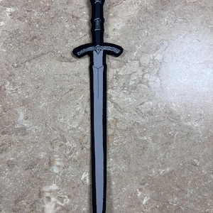 May include: A black and gray embroidered patch in the shape of a sword. The sword features a detailed hilt and crossguard, with a gray blade. The patch is likely designed for application on clothing or accessories.