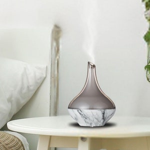 Peut inclure: Un diffuseur d'huiles essentielles en marbre blanc et gris avec un dessus gris et une base blanche. Le diffuseur est sur une table blanche et émet un panache de vapeur.