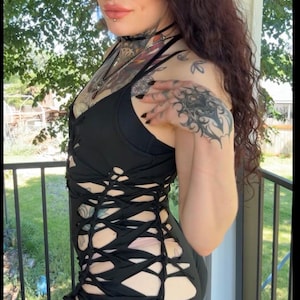 May include: A black mini dress with a unique cut-out design. The dress features a fitted bodice and a skirt with crisscross detailing. The model has dark hair and tattoos. The background includes a porch and greenery.