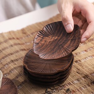 May include: A set of six brown wooden coasters with a unique leaf-like design. The coasters are stacked on top of each other, with the top coaster being held by a hand.