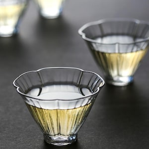 May include: Three clear glass teacups with fluted edges. The cups are filled with a light yellow liquid. The cups are sitting on a dark grey surface.