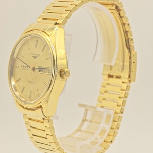 May include: A gold Longines Admiral automatic wristwatch. The watch has a gold dial with the day and date displayed. The watch has a gold strap and a gold crown. The watch is displayed on a clear stand.