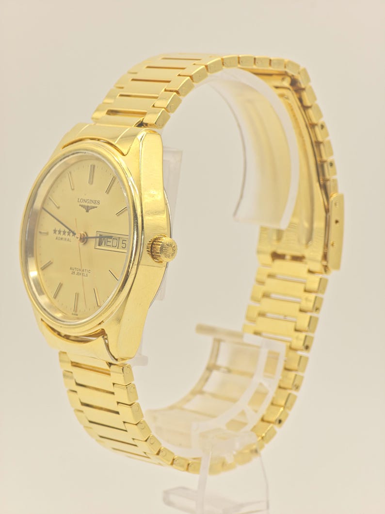 May include: A gold Longines Admiral automatic wristwatch. The watch has a gold dial with the day and date displayed. The watch has a gold strap and a gold crown. The watch is displayed on a clear stand.