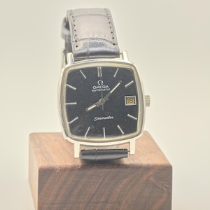 Könnte beinhalten: Eine Vintage Omega Seamaster Uhr mit einem schwarzen quadratischen Zifferblatt und einem Datumsfenster bei 3 Uhr. Die Uhr hat silberne Zeiger und Markierungen, ein silbernes Gehäuse und ein schwarzes Lederarmband. Die Uhr wird auf einem kleinen Holzblock präsentiert.