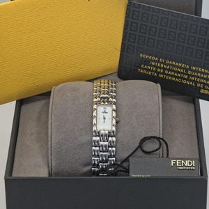 Può includere: Un orologio Fendi con cinturino argentato e quadrante bianco rettangolare, esposto in una scatola nera con interno grigio. L'orologio è accompagnato da una busta gialla e una carta nera con testo. L'orologio è Swiss Made.