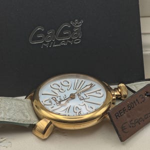May include: A gold GaGa Milano watch with a light blue face and large, stylized numerals. The watch has a light green leather strap and a textured crown. The watch is displayed with its black box, which has the GaGa Milano logo.