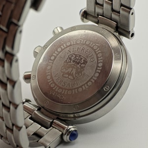May include: Close-up of a stainless steel Versace watch. The watch features a silver band and a round face with the text "ALFREDO VERSACE V414CH". The watch has blue accents on the band.