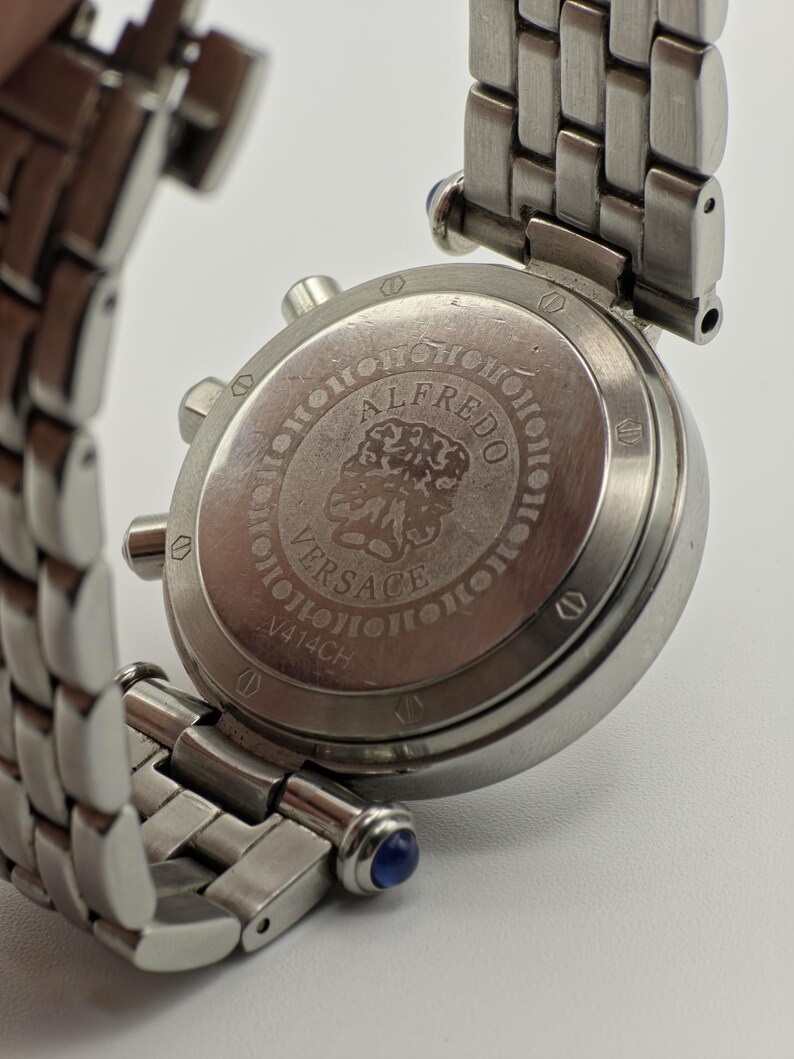 May include: Close-up of a stainless steel Versace watch. The watch features a silver band and a round face with the text "ALFREDO VERSACE V414CH". The watch has blue accents on the band.