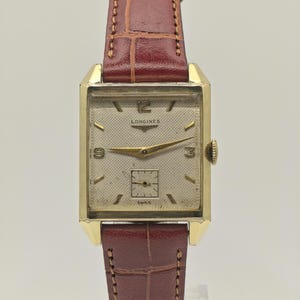 Könnte beinhalten: Eine Vintage-Longines-Armbanduhr mit einem rechteckigen, goldfarbenen Gehäuse und einem cremefarbenen Zifferblatt. Die Uhr verfügt über goldene Zeiger und Stundenmarkierungen, eine kleine Sekundenscheibe und ein braunes Lederarmband. Auf dem Zifferblatt steht "Longines" und "Swiss".
