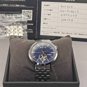 以下が含まれることがあります： 青い文字盤とシルバーのステンレススチールバンドを備えた、Bulovaの自動巻き腕時計。時計はオープンハートのデザインで、内部の仕組みを披露しています。黒い箱と書類に入っています。