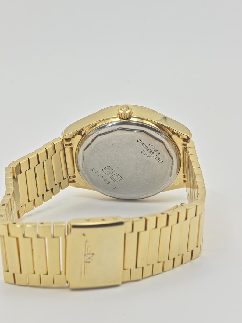 May include: A gold-coloured wristwatch with a stainless steel back. The watch has a rectangular link band and a clasp. The back of the watch has the text "STAINLESS STEEL BACK" and the number "27034503".