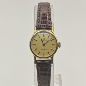 May include: An Omega De Ville wristwatch featuring a round gold-tone case and a champagne-colored dial with black hour markers. The watch includes a dark brown leather strap with a textured, embossed pattern and a small winding crown on the side.