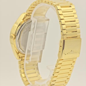 May include: Gold-coloured wristwatch with a rectangular face and a matching link bracelet. The watch features a stainless steel back and a clasp closure. The watch band is approximately 2 cm wide.