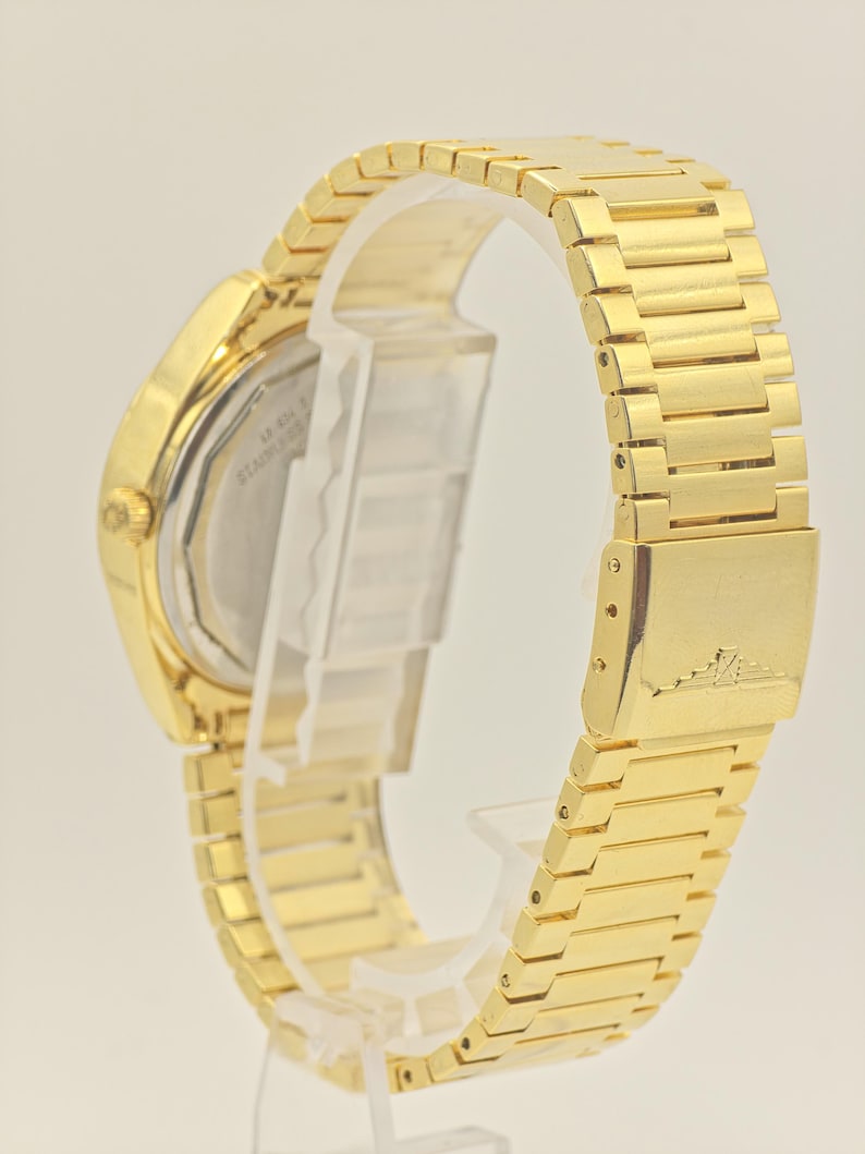 May include: Gold-coloured wristwatch with a rectangular face and a matching link bracelet. The watch features a stainless steel back and a clasp closure. The watch band is approximately 2 cm wide.