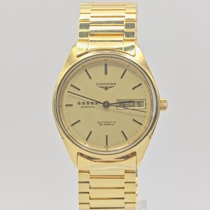 May include: A gold Longines Admiral automatic wristwatch. The watch has a gold dial with black hour markers and hands, a day-date display, and a gold link bracelet. The watch face displays the text "LONGINES", "Admiral", and "AUTOMATIC 25 JEWELS".