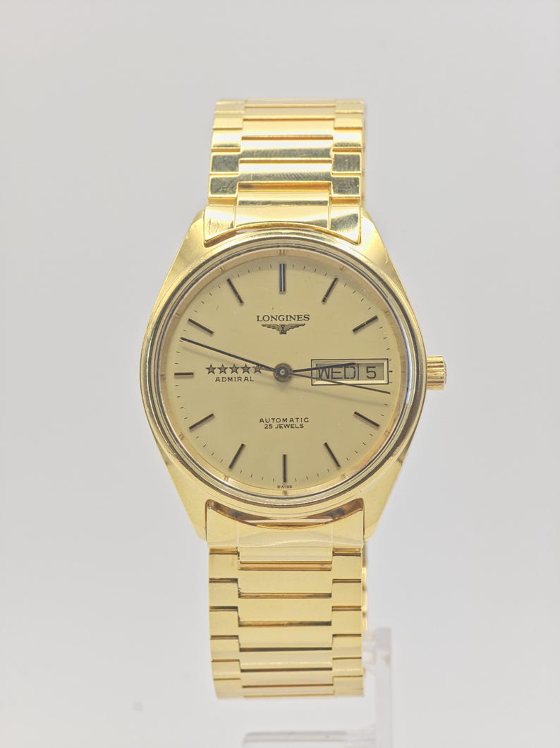 May include: A gold Longines Admiral automatic wristwatch. The watch has a gold dial with black hour markers and hands, a day-date display, and a gold link bracelet. The watch face displays the text "LONGINES", "Admiral", and "AUTOMATIC 25 JEWELS".