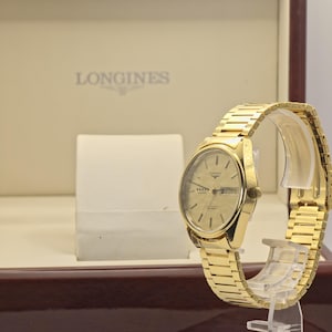 May include: A gold Longines quartz watch with a matching strap, displayed on a clear stand. The watch face has a gold dial with the Longines logo and the day of the week. The watch is presented in a brown box with the Longines logo.