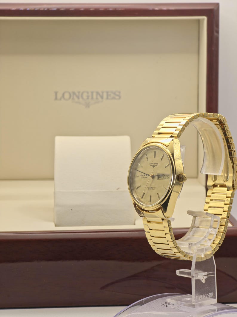 May include: A gold Longines quartz watch with a matching strap, displayed on a clear stand. The watch face has a gold dial with the Longines logo and the day of the week. The watch is presented in a brown box with the Longines logo.
