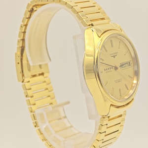 May include: A gold Longines Admiral automatic wristwatch. The watch has a gold dial with stick hour markers, a day-date window, and a gold bracelet. The watch is displayed on a clear stand.