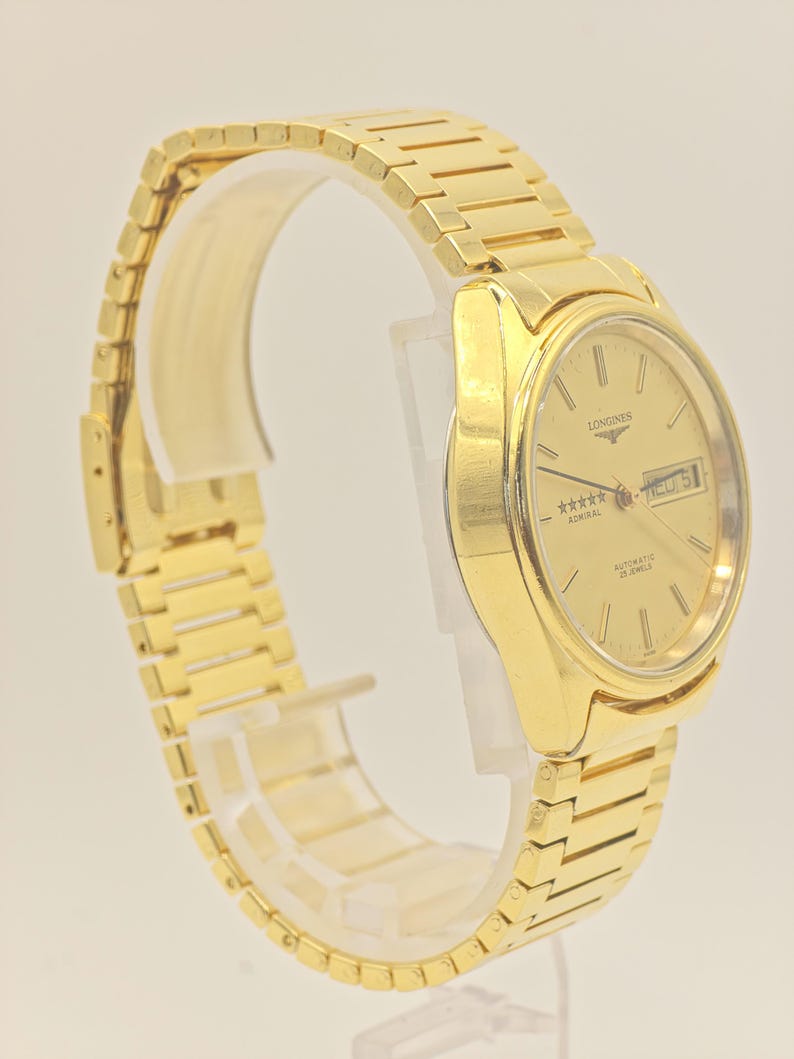 May include: A gold Longines Admiral automatic wristwatch. The watch has a gold dial with stick hour markers, a day-date window, and a gold bracelet. The watch is displayed on a clear stand.