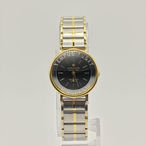 Pode incluir: Um relógio de quartzo Junghans dourado e prateado com mostrador preto. O relógio apresenta o texto "TEMPUS FUGIT" e "JUNGHANS Since 1861". A pulseira do relógio é uma combinação de elos prateados e dourados.