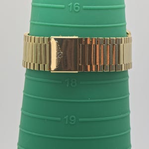 May include: A gold watch strap is shown on a green ring sizer. The watch strap is made of rectangular links and has a rectangular clasp with an emblem. The ring sizer has measurements from 16 to 20.