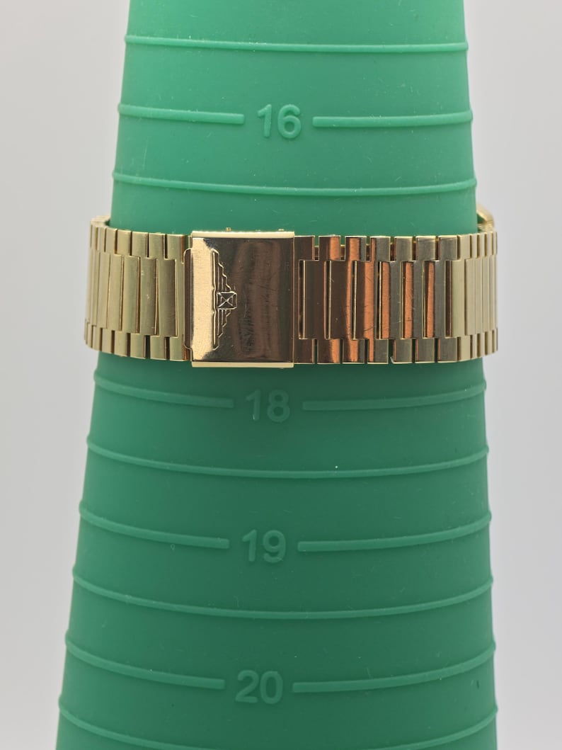 May include: A gold watch strap is shown on a green ring sizer. The watch strap is made of rectangular links and has a rectangular clasp with an emblem. The ring sizer has measurements from 16 to 20.