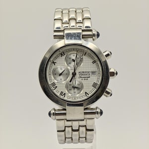 Może przedstawiać: Srebrny zegarek chronograf Alfredo Versace z białą tarczą i rzymskimi cyframi. Zegarek posiada wiele tarcz pomocniczych, wyświetlacz daty i bransoletę ze stali nierdzewnej. Zegarek ma niebieskie akcenty.