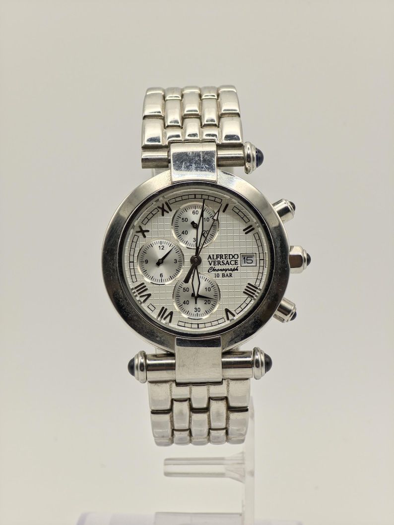 May include: A silver-toned Alfredo Versace chronograph watch with a white dial and Roman numeral hour markers. The watch features multiple sub-dials, a date display, and a stainless steel band. The watch has blue accents and is displayed on a clear stand.