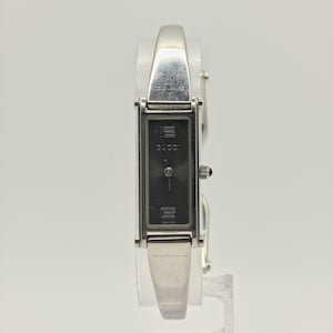 Puede incluir: Un reloj Gucci plateado con una esfera negra rectangular. El reloj tiene una correa plateada y una pequeña corona. El logotipo de Gucci es visible en la esfera. El reloj se exhibe sobre un soporte transparente.