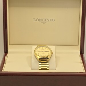 May include: A gold Longines watch with a gold strap, presented in a wooden box. The watch has a round face with a date display and is set against a cream-coloured interior. The box is made of dark, polished wood.