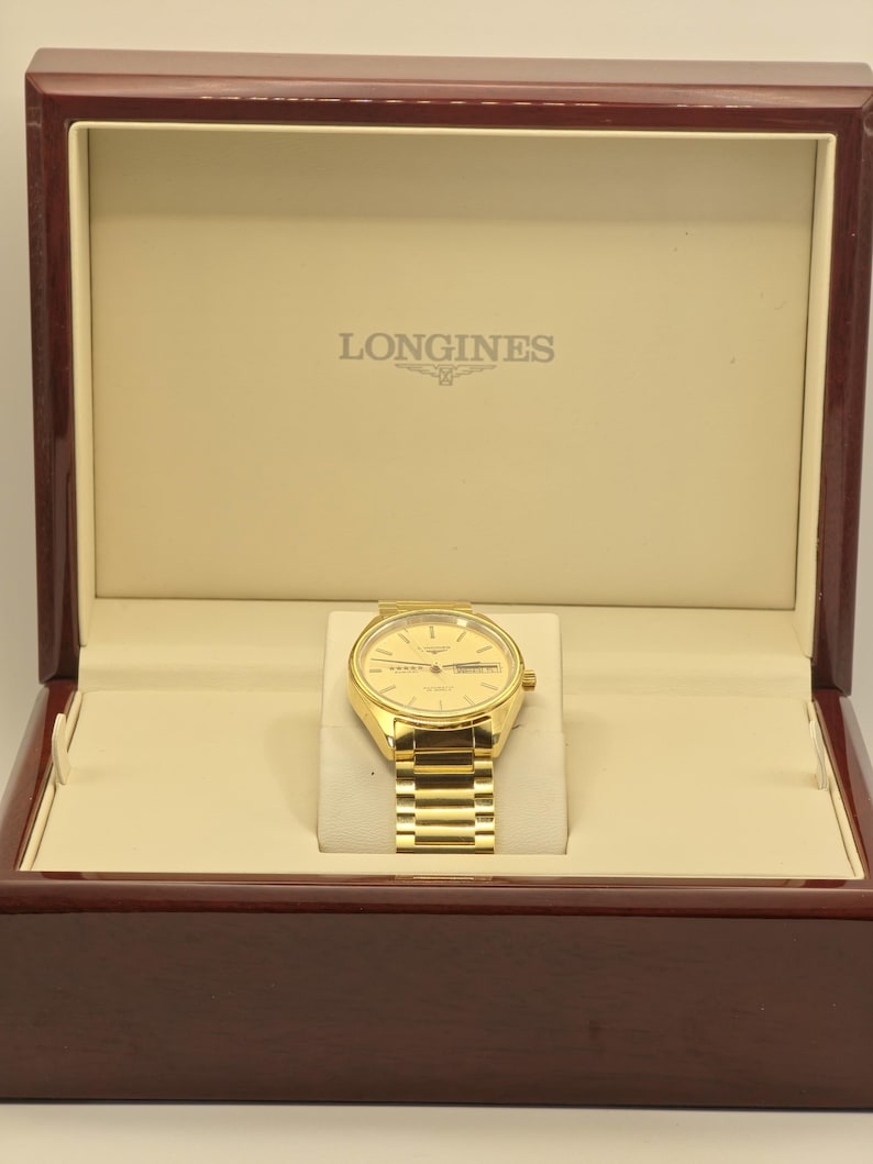 May include: A gold Longines watch with a gold strap, presented in a wooden box. The watch has a round face with a date display and is set against a cream-coloured interior. The box is made of dark, polished wood.