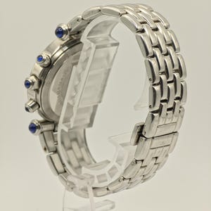 May include: A silver-toned wristwatch with a linked metal band. The watch face has three blue accents and a silver bezel. The watch is displayed on a clear plastic stand, showcasing the side profile of the watch.