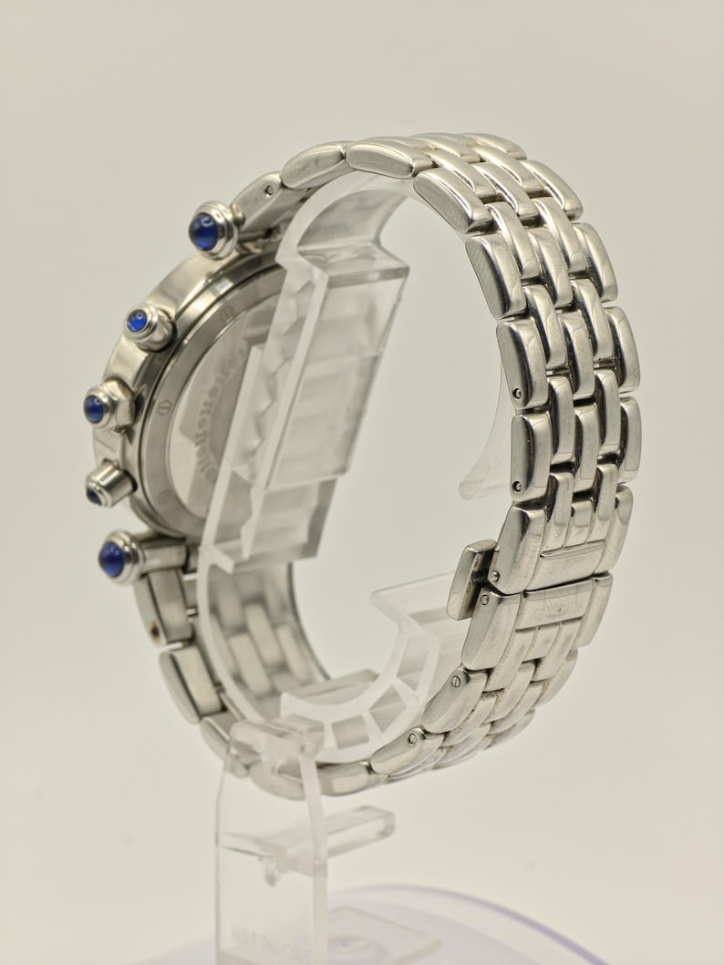 May include: A silver-toned wristwatch with a linked metal band. The watch face has three blue accents and a silver bezel. The watch is displayed on a clear plastic stand, showcasing the side profile of the watch.