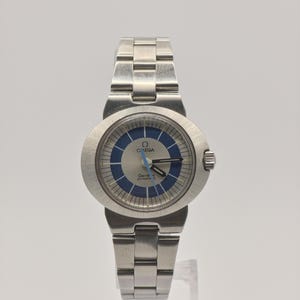Pode incluir: Um relógio de pulso Omega Geneve Dynamic prateado com mostrador azul e prateado. O relógio tem uma pulseira de aço inoxidável e um mostrador redondo. O relógio é exibido em um suporte transparente sobre um fundo branco.