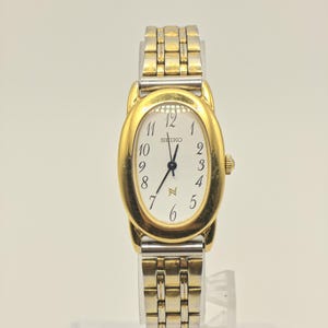 Può includere: Un orologio da polso Seiko con lunetta ovale color oro e quadrante bianco con numeri arabi neri. L'orologio presenta un cinturino a maglie in metallo bicolore argento e oro. Il quadrante mostra il marchio Seiko e un simbolo color oro.