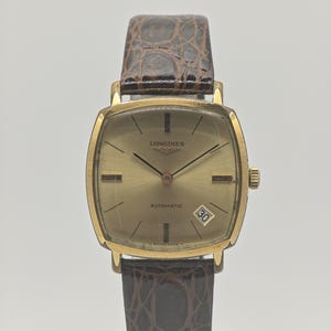 May include: A gold-toned Longines automatic wristwatch with a rectangular face and a brown leather band. The watch face displays the brand name, the word "AUTOMATIC", and a date window. The band has a crocodile pattern.