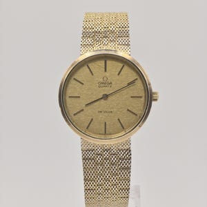 May include: A gold-toned Omega Quartz De Ville wristwatch. The watch features a round face with black hour markers and hands. The watchband is a woven gold mesh. The watch is displayed on a clear stand against a white background.