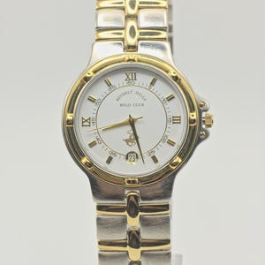 May include: A two-tone wristwatch with a silver and gold-colored metal link band. The round white dial features gold-tone hands, Roman numeral markers at twelve, three, and nine o'clock, a date window at six, and the text Beverly Hills Polo Club.
